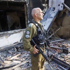 Man in military uniform stands in front of badly burned and damaged house