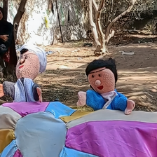 Two cloth puppets appear from behind a colorful piece of cloth on a sunny day.