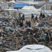Piles of trash make people walking by look tiny, tents in the background