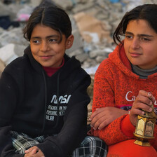 Two young girls sit on rubble, one holding a small lantern
