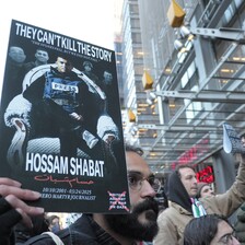 Man holds poster showing Hossam Shabat in press jacket sitting in armchair