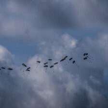 A backdrop of clouds and small black packages with parachutes attached being dropped from the back of an airplane.