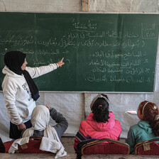 A teacher in a tent points to a blackboard as she stands in front of several children 