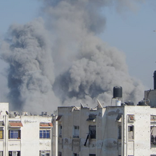 In daytime, massive clouds of black-gray smoke appear over city buildings