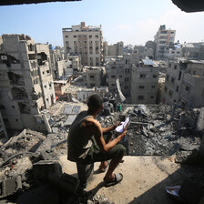 A young man holding a notebook sits on a chair overlooking a cityscape  of destroyed buildings