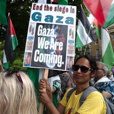 Woman wearing sunglasses holds picket sign reading: "End the siege in Gaza. Gaza, we're coming."