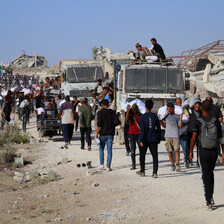People walk alongside trucks on a dirt road in the daytime, carrying aid. 
