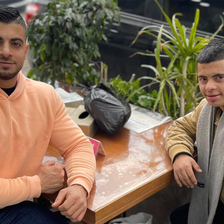 Two men sit outdoors with their elbows resting on a table