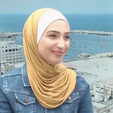 Young woman wearing a jean jacket and mustard yellow hijab smiles slightly while sitting in front of backdrop of Gaza port