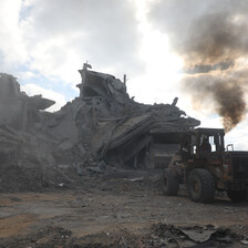 A bulldozer clears rubble from a destroyed house with smoke still billowing in the background