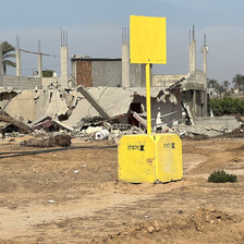 A concrete block painted yellow with a yellow sign stands before the rubble of buildings