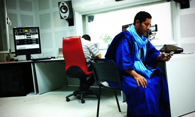 Man wearing blue robe looks at his phone in a radio station suite