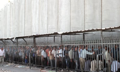 Palestinian men queue at checkpoint next to very tall concrete wall