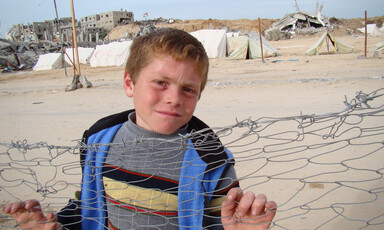 Boy leans against fence with destroyed homes in background