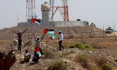 Youths holding Palestinian flags stand in front of Israel's border wall and military jeep