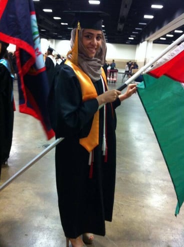 Young woman wearing graduation gown smiles while holding Palestinian flag