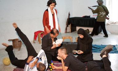 Scene from play shows family sitting together in foreground with Israeli soldier character in background