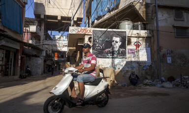 Scene shows youth on scooter riding past wall covered with poster and graffiti