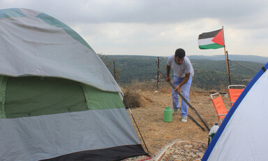 Man picks up bar amid tents with Palestine flag and landscape in background