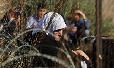 Elderly woman sits next to suitcase near fence and barbed wire