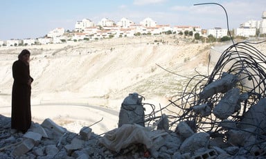 Palestinian woman stands among rubble of destroyed home with Israeli settlement in background