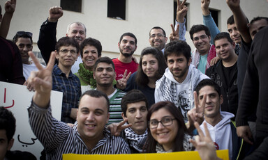 Young man smiles among crowd making victory symbol with their hands