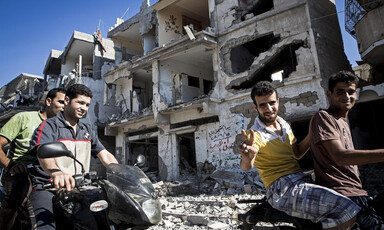 Smiling young men on motorbikes give victory sign in front of shelled building in Gaza City