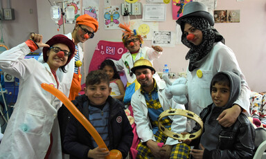 Men and women in silly hats and wigs wearing red foam noses pose with children in hospital room