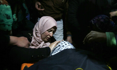 Crying woman places hand on forehead of body wrapped in black flag and checkered scarf