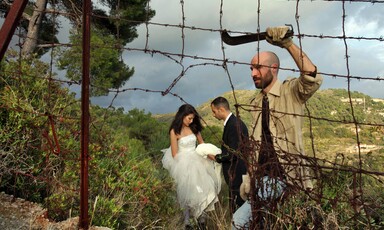 Man with machete makes hole in barbed wire fence as couple dressed as bride and groom stand in background in natural landscape