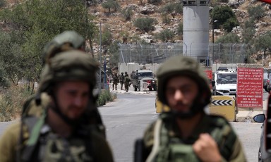 Israeli soldiers stand in foreground with military checkpoint watchtower and crowd of people in background