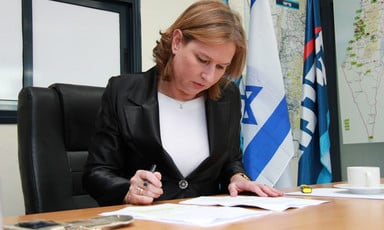Tzipi Livni sits at desk looking at paper with an Israeli flag hanging behind her