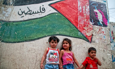 Small children stand against wall with mural of Palestine flag