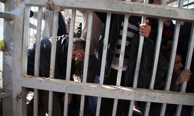 Crowd of men stand or sit behind metal bars at checkpoint