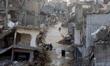 View of people walking through muddy road in between bombed-out buildings