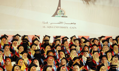 Women students wearing graduation caps and gowns sit in front of Al Aqsa University banner