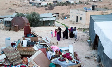 Women and children stand near home with possessions piled up outside