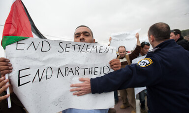 Israeli police officer places arm across Palestinian protester carrying sign reading End Settlements and Apartheid