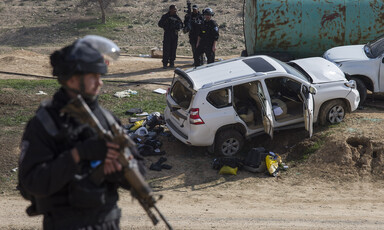 Militarized police officer holding rifle stands in foreground with SUV emptied of its contents in background