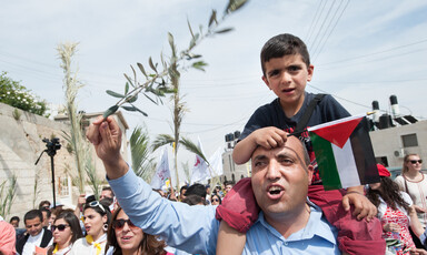 Man waving olive branch carries boy holding Palestine flag on his shoulders during Palm Sunday procession