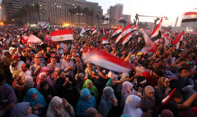 Crowd of hundreds of men and women wave Egyptian flags in public square
