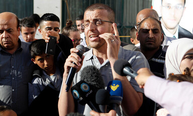 Man talks into microphone as crowd of people stand around him
