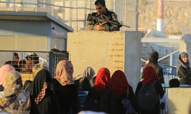 Palestinian woman walk through a checkpoint as an armed Israeli soldier is positioned above them