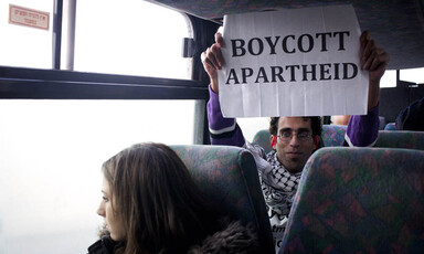 Basil al-Araj sits on bus behind Israeli woman while holding up a sign reading Boycott Apartheid