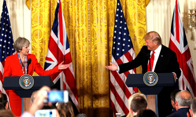 Theresa May and Donald Trump, standing at podiums during White House press conference, smile and reach their arms towards one another