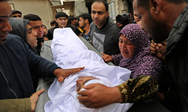 Woman uses her hand to cup the head of teen wrapped in burial shroud as crowd gathers around them