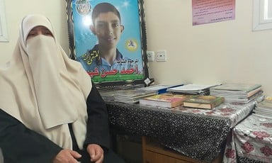 Woman with covered face sits next to table and poster of her smiling teenage son