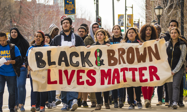 Racially diverse group of young people carry banner reading Black and Brown Lives Matter 