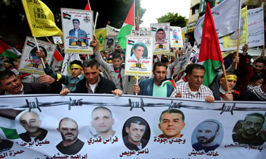 Group of men hold banner showing images of jailed Palestinians in front of crowd of people holding up Palestinian flags and posters of prisoners