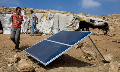 A boy stands next to a solar panel installed on barren ground with makeshift shelters in the background
